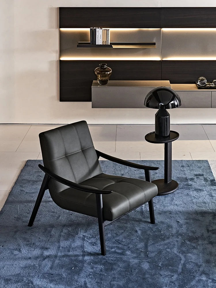 A modern black Marc leather lounge chair with a tufted backrest and wooden legs, positioned on a textured blue rug next to a black side table with a dome lamp, in front of a contemporary dark wood and light gray wall cabinet.