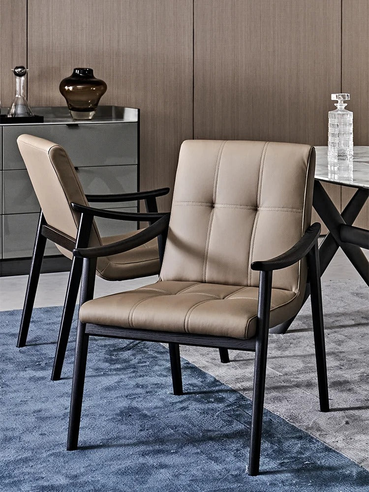 A modern upholstered dining chair with beige tufted leather and a black metal frame, shown in a styled interior setting with a second matching chair, a cabinet, and a blue rug.