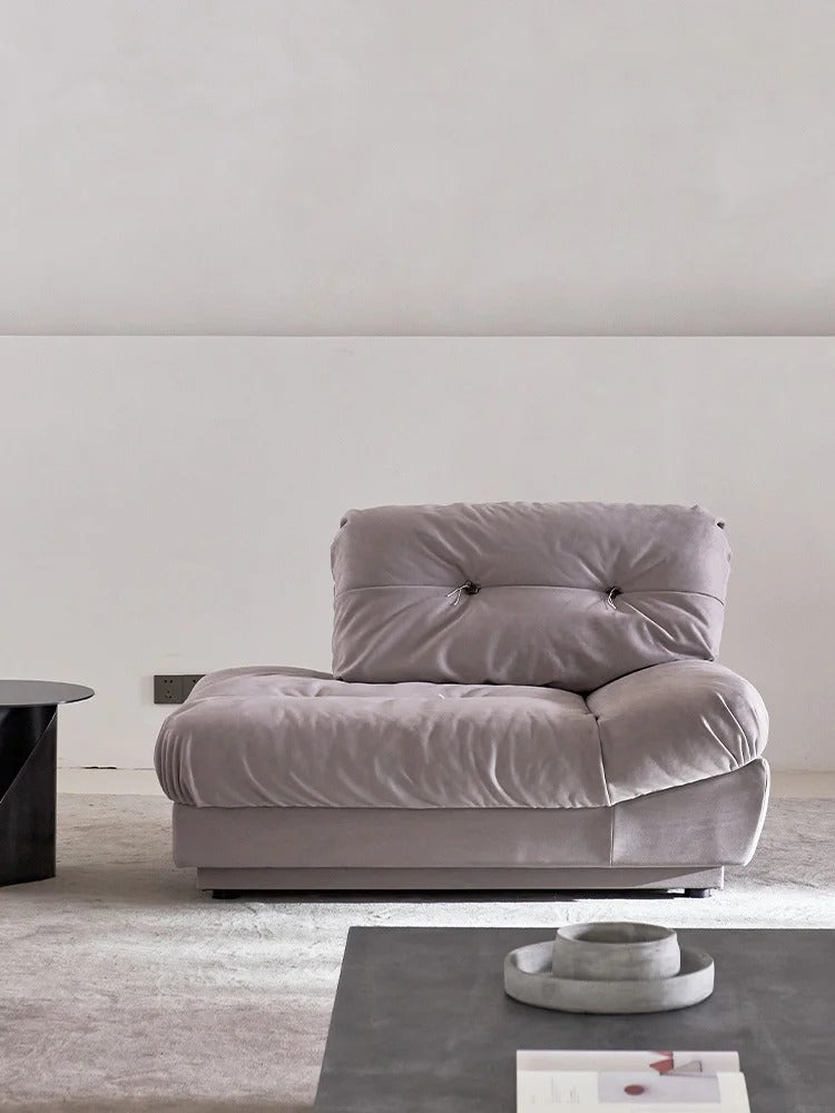 A modern tufted lounge chair in a dusty lavender-gray velvet fabric, set in a minimalist living room with a dark coffee table and textured wall.
